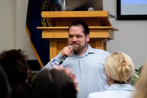 Tim Houle, in a light blue shirt stands in front of a wooden podium, holding a microphone and speaking to an audience. He is smiling and looking to the side. Behind him is a flag and a projection screen. Several people in the foreground are facing him, listening attentively.