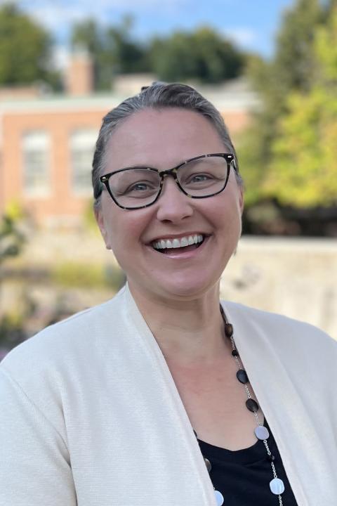 Heidi Cloutier is a woman salt-and-pepper hair pulled back, wearing black rimmed glasses, a black top, and white jacket. She is standing outdoors on a sunny day and smiling